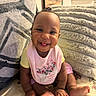 baby, smiling, sitting, indoors, blanket, cushion, cozy, happy, child, pink_bib, yellow_shirt, cute, portrait, home, infant, foot, hand, skin, hair_tie, expression