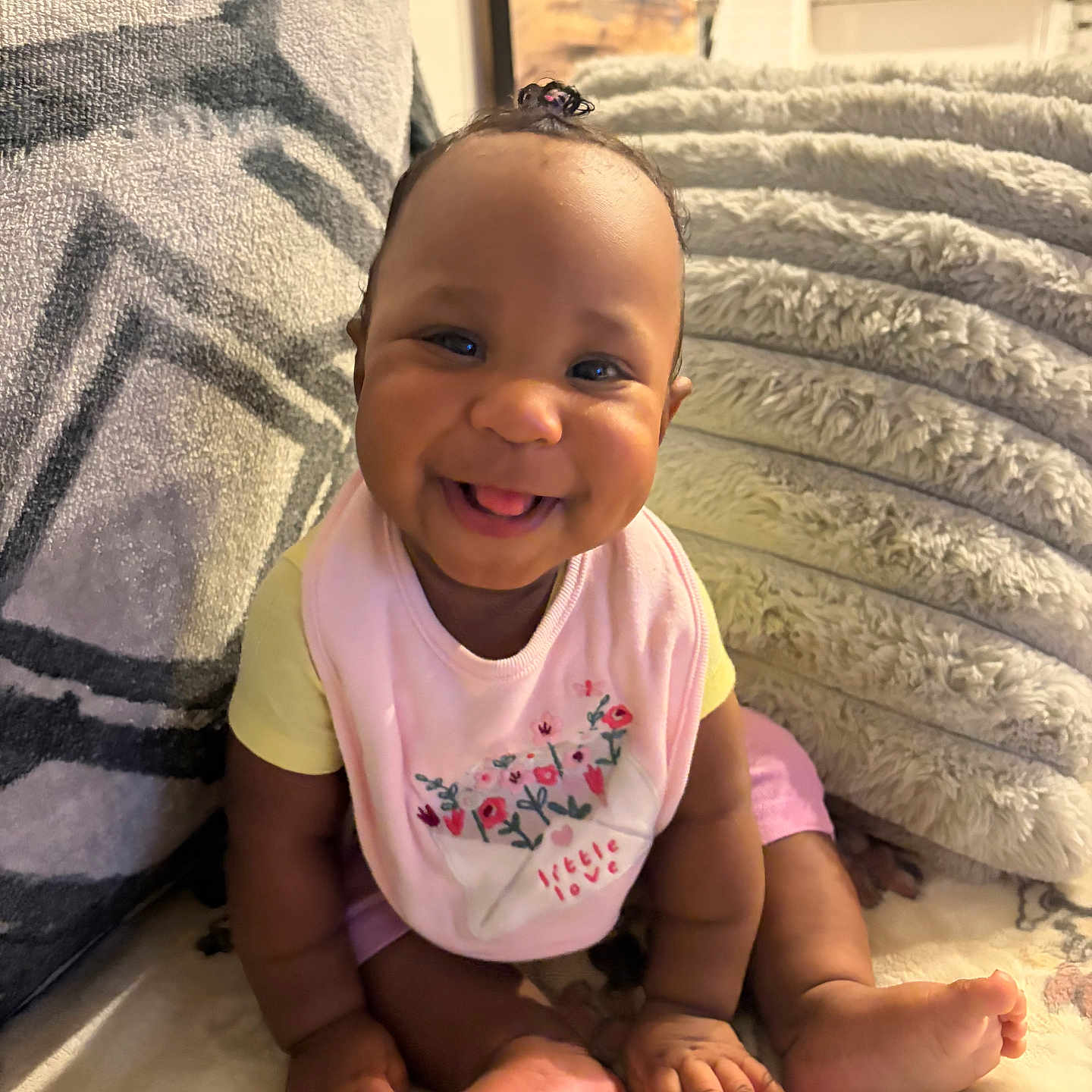 Emmani joined the competition — help win amazing prizes! baby, smiling, sitting, indoors, blanket, cushion, cozy, happy, child, pink_bib, yellow_shirt, cute, portrait, home, infant, foot, hand, skin, hair_tie, expression