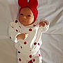 baby, infant, red_bow, white_outfit, apple_prints, red_socks, lying_down, blanket, cute, newborn, child, portrait, indoors, clothing, headwear, fist, expression, soft_light, fabric, resting