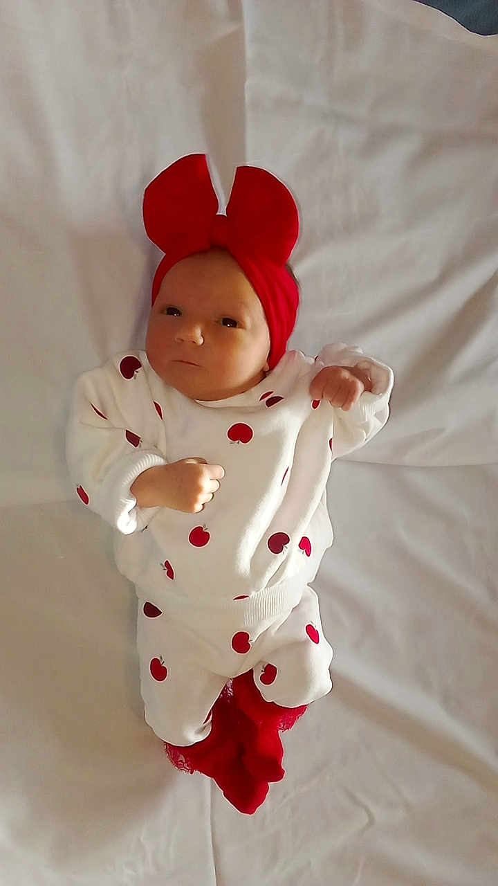 Thalya participe au concours pour gagner de l'argent avec cette photo : baby, infant, red_bow, white_outfit, apple_prints, red_socks, lying_down, blanket, cute, newborn, child, portrait, indoors, clothing, headwear, fist, expression, soft_light, fabric, resting
