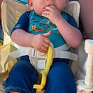 Diego a rejoint le concours — aidez-le/la à gagner de superbes lots ! baby, child, blue_clothing, high_chair, yellow_toy, hand, face, person, sitting, fingers_in_mouth, short_hair, sunlight, outdoor, adult, seatbelt, legs, foot, skin, expression, toddler