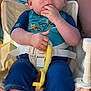baby, child, blue_clothing, high_chair, yellow_toy, hand, face, person, sitting, fingers_in_mouth, short_hair, sunlight, outdoor, adult, seatbelt, legs, foot, skin, expression, toddler