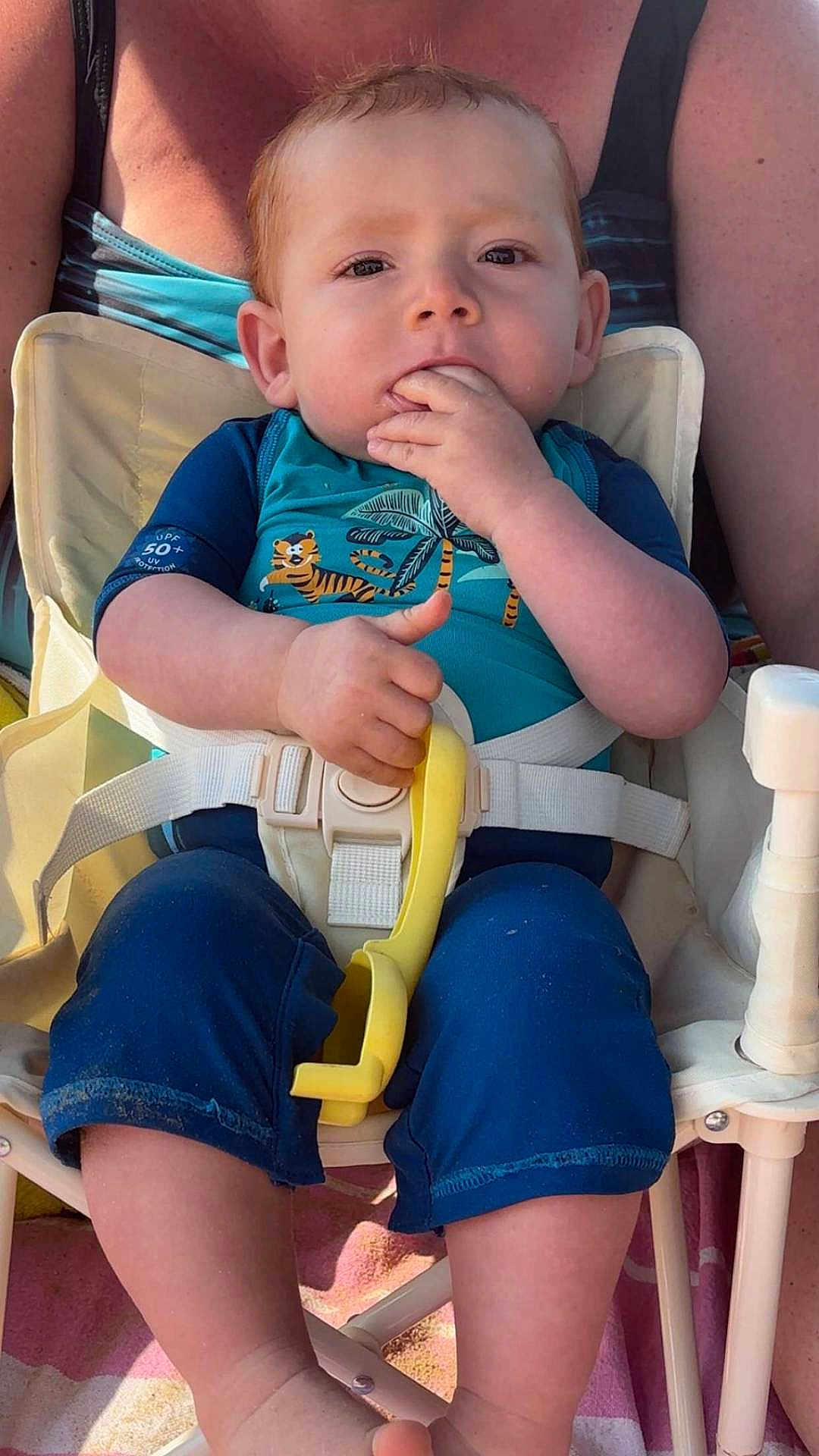 Diego a rejoint le concours — aidez-le/la à gagner de superbes lots ! baby, child, blue_clothing, high_chair, yellow_toy, hand, face, person, sitting, fingers_in_mouth, short_hair, sunlight, outdoor, adult, seatbelt, legs, foot, skin, expression, toddler