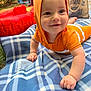 baby, child, orange_clothing, hood, blanket, plaid, outdoor, grass, drool, hand, smile, face, toy, inflatable, tote_bag, curious, crawling, playful, sunlight, happy