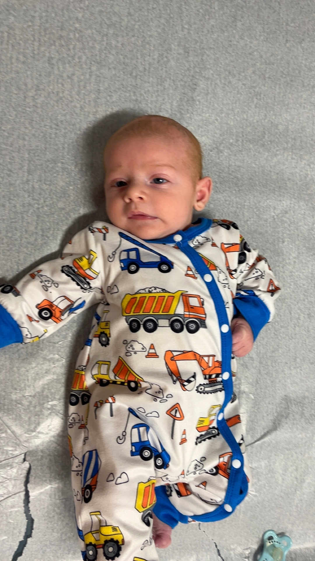 Blake is registered to the contest to win money with this photo: baby, infant, onesie, construction_theme, blue_trim, pacifier, lying_down, gray_background, cute, clothing, child, face, small_hands, foot, soft_surface, indoor, expression, patterned_clothing, newborn, resting