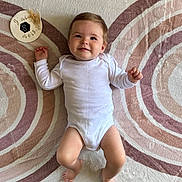 Emma a rejoint le concours — aidez-le/la à gagner de superbes lots ! baby, infant, child, onesie, smile, face, lying_down, rug, floor, milestone, decor, pattern, concentric_circles, plush, skin, hands, feet, head, happy, portrait