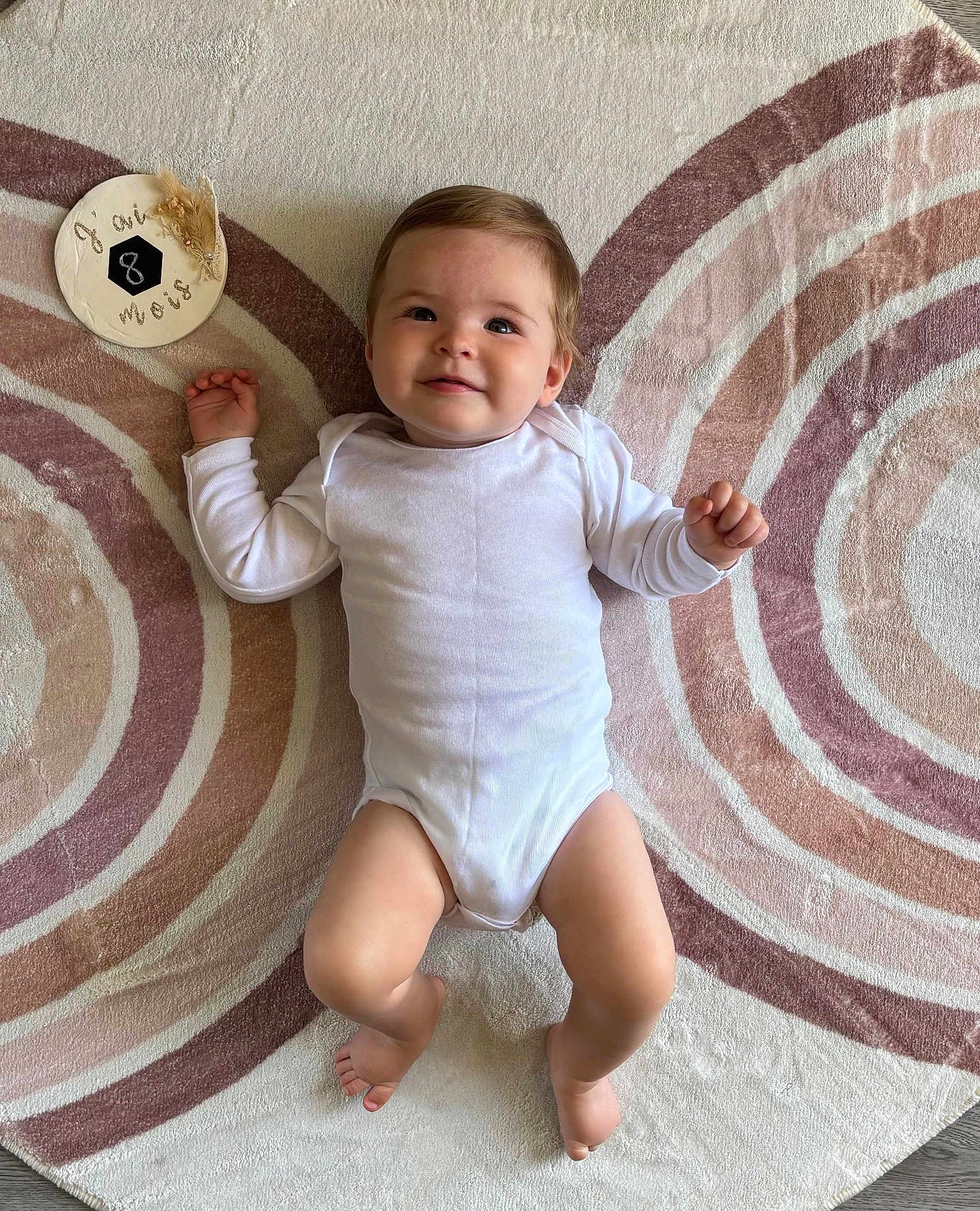 Emma a rejoint le concours — aidez-le/la à gagner de superbes lots ! baby, infant, child, onesie, smile, face, lying_down, rug, floor, milestone, decor, pattern, concentric_circles, plush, skin, hands, feet, head, happy, portrait