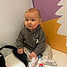 baby, child, indoor, toy, striped_pants, hoodie, sitting, curious, playful_background, white_surface, black_strap, colorful_toy, infant, cute, young_child, casual_clothing, portrait, small_toy, expression, light