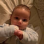 baby, infant, child, face, big_eyes, hand_in_mouth, onesie, blanket, bedding, bed, pillow, portrait, close_up, indoors, soft_lighting, cute, sleepwear, small_hands, gazing, smile
