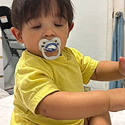 Martinez is registered to the contest to win money with this photo: toddler, baby, pacifier, yellow_shirt, diaper, sitting, indoors, child, sleepy, cute, person, foot, arm, face, hair, television, furniture, room, household, casual