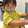 toddler, baby, pacifier, yellow_shirt, diaper, sitting, indoors, child, sleepy, cute, person, foot, arm, face, hair, television, furniture, room, household, casual