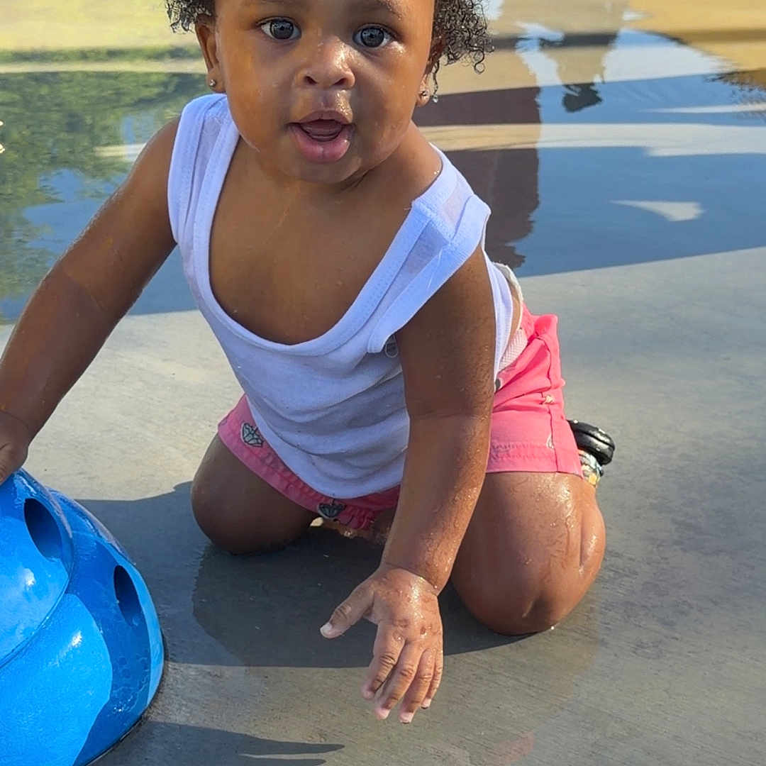 Maziyon is registered to the contest to win money with this photo: toddler, child, water, playground, splash_pad, blue, white_tank_top, pink_shorts, wet_surface, outdoor, sunlight, reflection, shadow, curious, kneeling, play, fun, summer, water_play, person