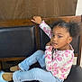 child, girl, curly_hair, pigtails, sitting, bench, black_cushion, pink_butterfly_shirt, light_blue_jeans, brown_shoes, bracelet, earrings, necklace, indoor, wooden_bench, casual_clothing, portrait, looking_at_camera, young, relaxed