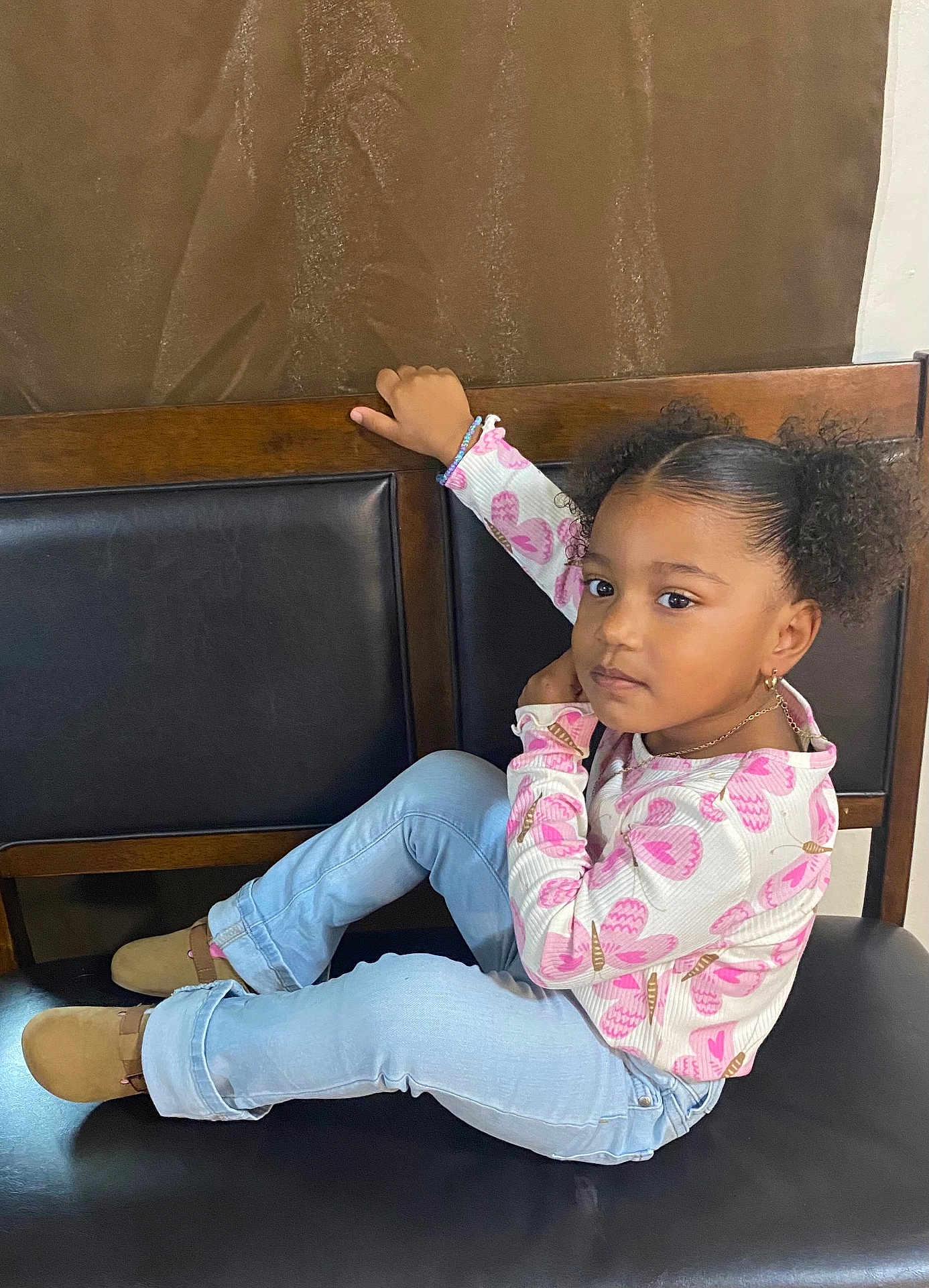 Noelie joined the competition — help win amazing prizes! child, girl, curly_hair, pigtails, sitting, bench, black_cushion, pink_butterfly_shirt, light_blue_jeans, brown_shoes, bracelet, earrings, necklace, indoor, wooden_bench, casual_clothing, portrait, looking_at_camera, young, relaxed