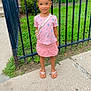 child, girl, sidewalk, fence, iron_fence, greenery, grass, bushes, pink_clothing, skirt, floral_pattern, sandals, hair_bun, smiling, outdoor, daylight, concrete, person, small_purse, casual