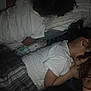child, children, sleeping, resting, bed, pillow, blanket, white_shirt, curly_hair, indoor, dim_light, adult, glasses, face_partial, close_together, dark, grainy, casual, family, home