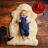 baby, infant, denim_overalls, smiling, face, fluffy_rug, wooden_floor, cowboy_boots, cowboy_hat, footwear, hat, child, portrait, happy, indoor, clothing, person, cute, lying_down, studio_photo