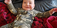 Anthony is registered to the contest to win money with this photo: baby, child, smiling, camo_pajamas, leather_couch, blanket, red_blanket, plaid_blanket, happy, indoors, person, infant, face, cozy, home, furniture, cute, relaxed, sitting, portrait