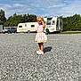 child, girl, pink_dress, pink_shoes, gravel, outdoor, sunny, daylight, car, trailer, camper, trees, sky, clouds, playing, standing, shadow, smiling, happy, nature