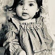 Ariella is registered to the contest to win money with this photo: baby, child, portrait, black_and_white, sitting, dress, shoe, blanket, pattern, indoor, cute, hair_bow, face, expression, infant, young_child, person, texture, closeup, monochrome