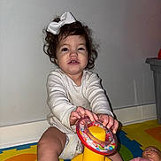 Carlotta is registered to the contest to win money with this photo: toddler, child, toy, doll, play_mat, white_bow, curly_hair, indoor, diaper, long_sleeve_shirt, colorful, plastic_toy, floor, baby, cute, sitting, playtime, person, young_child, smiling