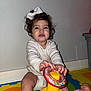 toddler, child, toy, doll, play_mat, white_bow, curly_hair, indoor, diaper, long_sleeve_shirt, colorful, plastic_toy, floor, baby, cute, sitting, playtime, person, young_child, smiling