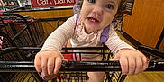 Carlotta is registered to the contest to win money with this photo: toddler, child, shopping_cart, store, floor, hand, face, shoes, clothing, necklace, person, indoor, wood_floor, retail, curly_hair, smile, shopping, toy, girl, baby