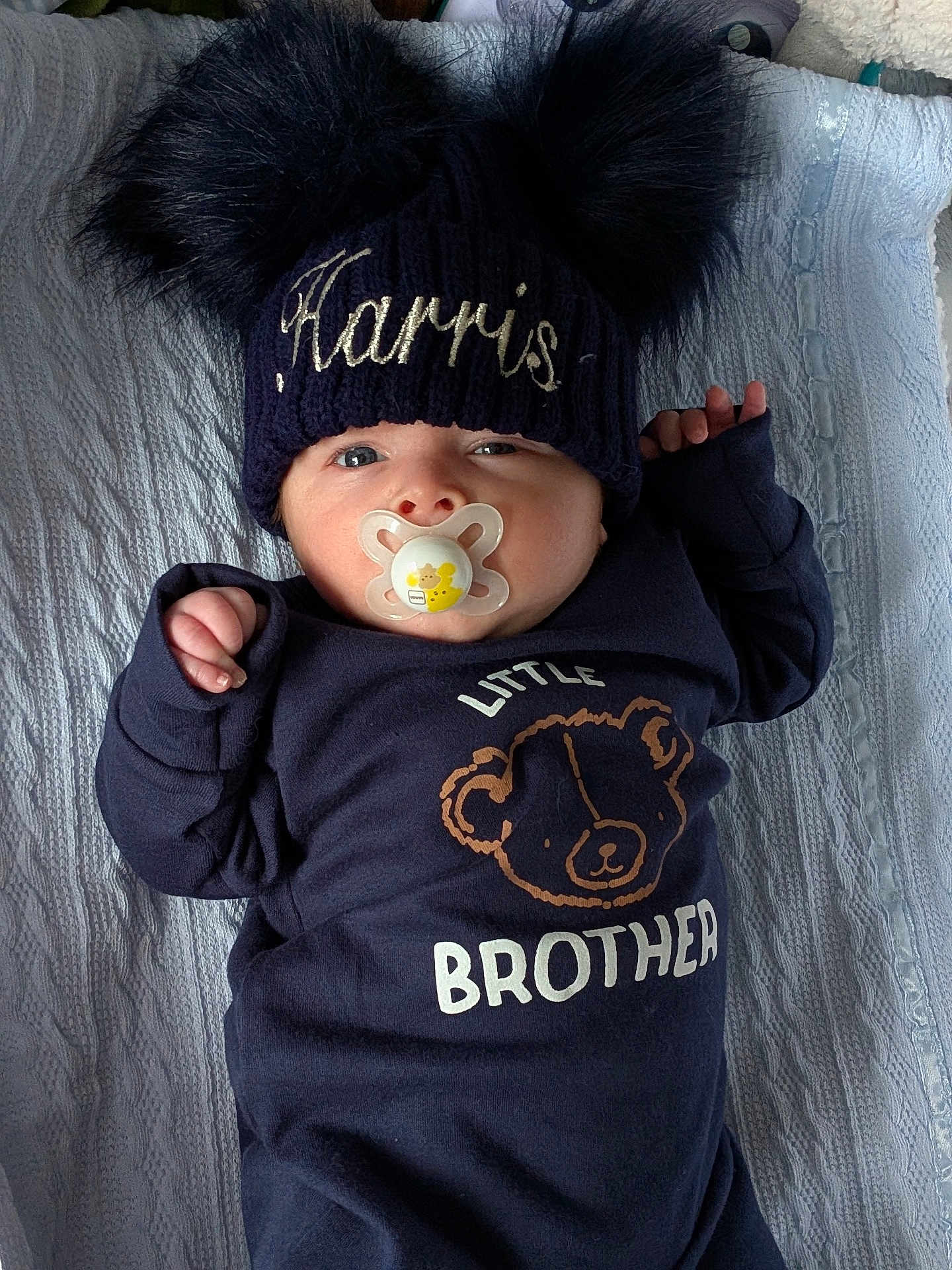 Harris joined the competition — help win amazing prizes! baby, pacifier, hat, pom_poms, knit_hat, blue_clothing, bear_graphic, text_on_clothing, infant, blanket, lying_down, close_up, cute, child, face, expression, indoor, portrait, newborn, sleeper