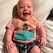 Luca is registered to the contest to win money with this photo: baby, infant, smiling, happy, tshirt, patterned_shorts, cushion, sitting, indoor, cute, child, portrait, skin, hand, legs, head, person, clothing, expression, comfort
