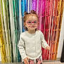 toddler, child, glasses, pink_glasses, white_cardigan, floral_skirt, smiling, happy, standing, indoor, rainbow_backdrop, colorful, metallic_fringe, hair_buns, cute, portrait, floor_tiles, playful, fashion, person