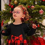Naomy participe au concours pour gagner de l'argent avec cette photo : child, toddler, glasses, dress, bow, red, black, checkered, christmas_tree, ornaments, holiday, curly_hair, headband, indoor, smiling, festive, person, celebration, happy, decorations