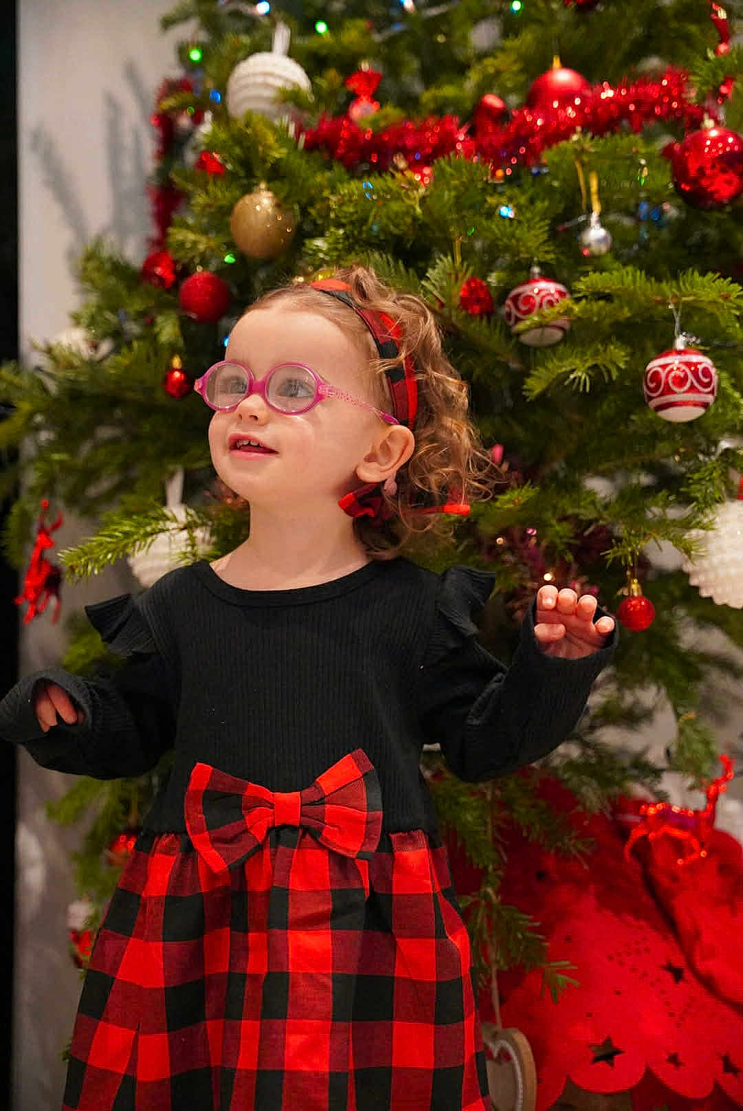 Naomy participe au concours pour gagner de l'argent avec cette photo : child, toddler, glasses, dress, bow, red, black, checkered, christmas_tree, ornaments, holiday, curly_hair, headband, indoor, smiling, festive, person, celebration, happy, decorations