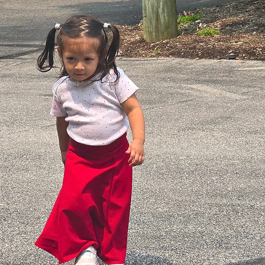 Isiani Elizabeth joined the competition — help win amazing prizes! asphalt, bushes, casual_clothing, child, curly_hair, daylight, fence, girl, greenery, hair_ties, nature, outdoor, person, pigtails, pole, red_skirt, shoes, sidewalk, walking, white_shirt