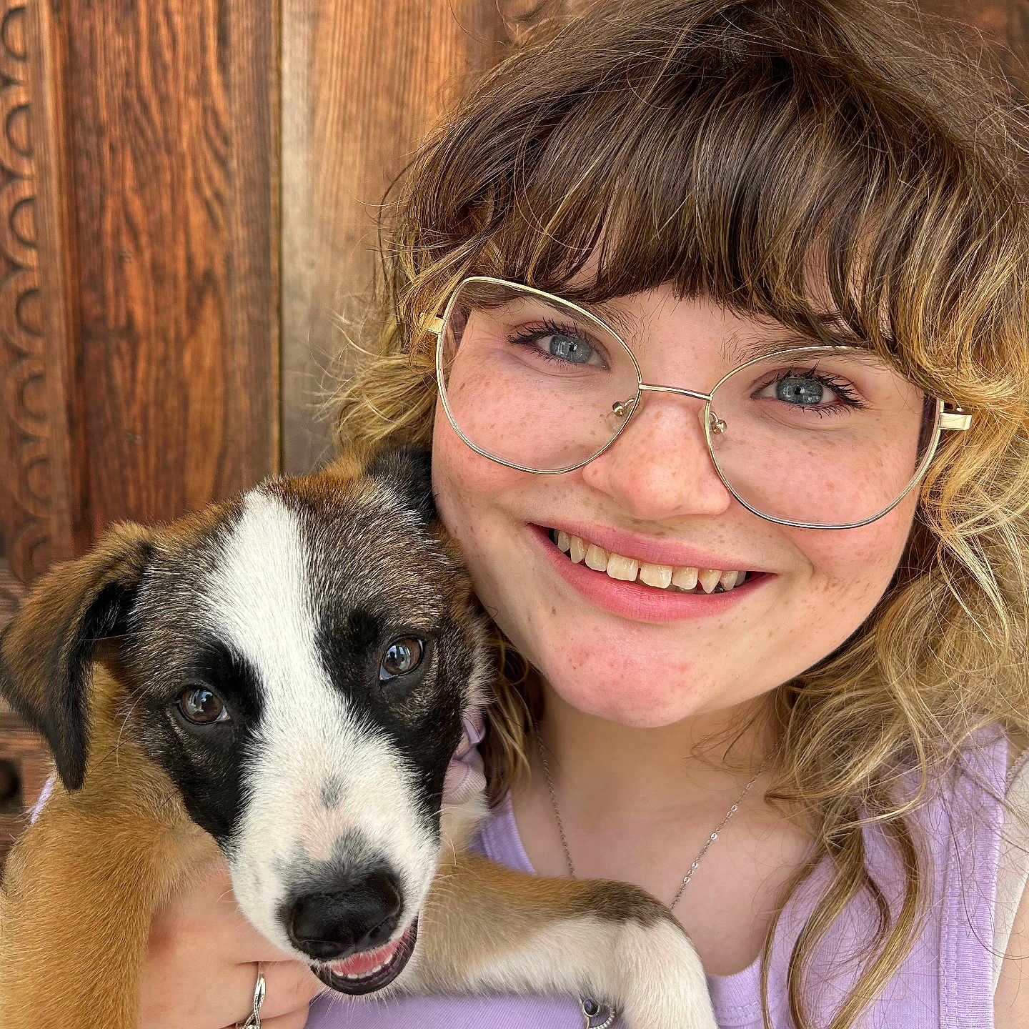 Maxinne joined the competition — help win amazing prizes! casual_clothing, closeup, curly_hair, dog, face, freckles, friendly, glasses, happy, holding, jewelry, light_purple_top, outdoor, pet, portrait, puppy, smile, woman, wooden_door, young_adult