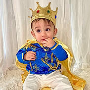 Thiago is registered to the contest to win money with this photo: toddler, child, crown, costume, blue_jacket, gold_embroidery, cape, white_pants, socks, indoor, curtains, fluffy_rug, portrait, cute, baby, seated, royalty, playful, fashion, smiling