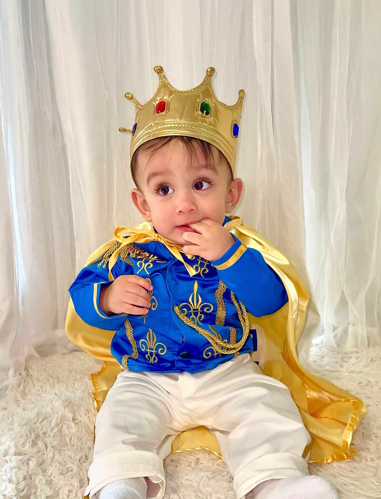 Thiago is registered to the contest to win money with this photo: toddler, child, crown, costume, blue_jacket, gold_embroidery, cape, white_pants, socks, indoor, curtains, fluffy_rug, portrait, cute, baby, seated, royalty, playful, fashion, smiling