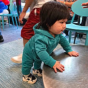 Ryan Mavo is registered to the contest to win money with this photo: child, toddler, baby, indoor, party, balloons, chairs, table, playroom, teal_clothing, tracksuit, sneakers, standing, exploring, hands, brown_hair, profile, adults, birthday, carpet