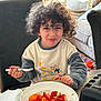 child, couch, curly_hair, cushion, dining, food, fork, fruit, happy, indoor, living_room, meal, natural_light, orange_fruit, person, plate, smile, strawberries, sweatshirt, table