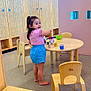 child, toddler, girl, barefoot, pink_shirt, blue_shorts, wooden_table, toy_dishes, playroom, classroom, wooden_chairs, cubby_storage, smiling, indoor, flooring, furniture, happy, person, young_child, play