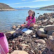 Lucia is registered to the contest to win money with this photo: child, girl, lake, rocks, water, sky, mountains, sunlight, pink_clothing, minnie_mouse, outdoor, nature, play, sitting, funny_face, container, bag, daylight, recreation, scenic