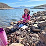 child, girl, lake, rocks, water, sky, mountains, sunlight, pink_clothing, minnie_mouse, outdoor, nature, play, sitting, funny_face, container, bag, daylight, recreation, scenic
