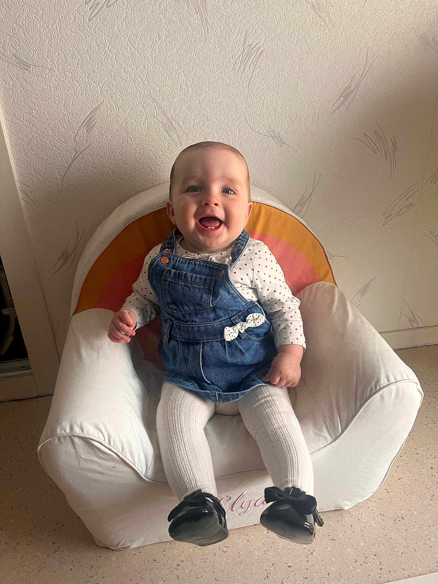 Elya participe au concours pour gagner de l'argent avec cette photo : baby, child, smiling, denim_dress, white_tights, black_shoes, armchair, rainbow_pattern, indoor, happy, cute, sitting, person, face, head, furniture, floor, wall, home, cozy
