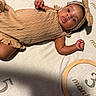 baby, infant, child, portrait, lying_down, blanket, tan_outfit, headband, bow, textured_clothing, wide_eyes, tongue_out, face, arms, legs, indoor, bed, soft_lighting, patterned_background, photography