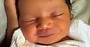 Jazaria is registered to the contest to win money with this photo: baby, newborn, sleeping, smiling, portrait, close_up, face, infant, white_clothing, swaddled, indoors, bed, bedding, hand, cozy, peaceful, soft_lighting, dark_hair, relaxed, family