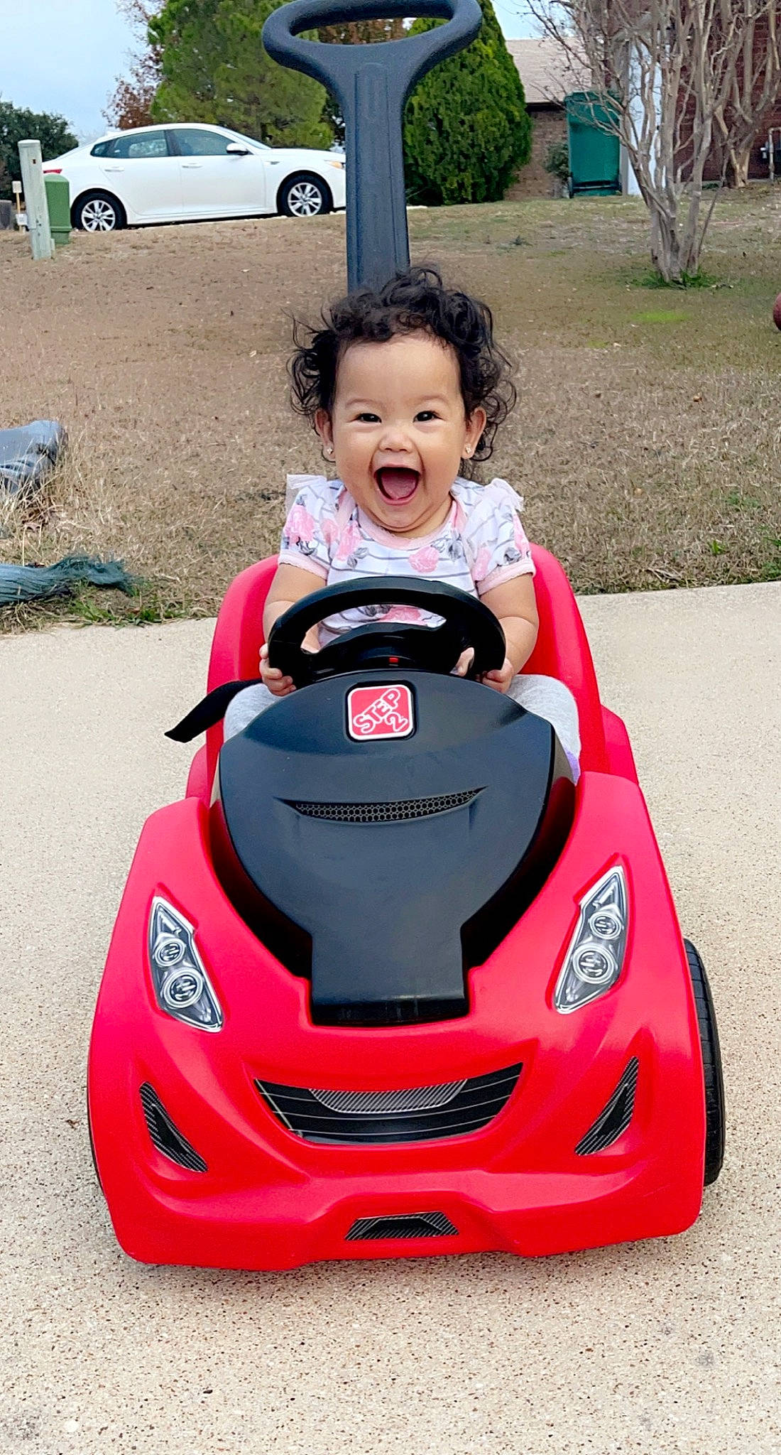 Aleila is registered to the contest to win money with this photo: automotive_design, automotive_exterior, automotive_lighting, automotive_tire, bumper, car, fender, grass, motor_vehicle, person, photograph, pink, plant, red, riding_toy, smile, tire, toddler, vehicle, wheel