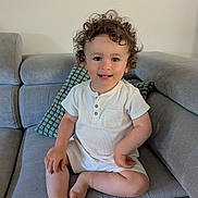 Arthur participe au concours pour gagner de l'argent avec cette photo : toddler, child, curly_hair, smiling, white_clothing, romper, couch, gray_couch, pillow, indoor, seated, barefoot, cute, happy, portrait, home, soft_lighting, young_child, face, person
