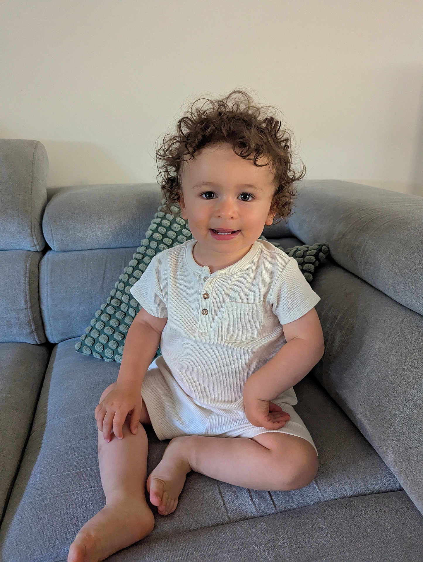 Arthur participe au concours pour gagner de l'argent avec cette photo : toddler, child, curly_hair, smiling, white_clothing, romper, couch, gray_couch, pillow, indoor, seated, barefoot, cute, happy, portrait, home, soft_lighting, young_child, face, person