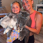 Moto Moto is registered to the contest to win money with this photo: woman, child, cat, indoor, smiling, happy, fluffy_cat, holding, living_room, floor, casual_clothing, bracelet, curtains, wall, candle, shelf, wood_floor, kneeled, portrait, family