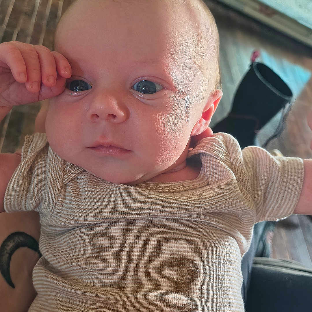 Noah is registered to the contest to win money with this photo: baby, blurred_background, child, curious, cute, expression, eyes, face, floor, hand, indoor, infant, newborn, onesie, person, portrait, skin, striped_clothing, wooden_floor, young