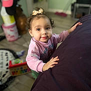 Stormie is registered to the contest to win money with this photo: toddler, child, indoor, curly_hair, pink_bow, pink_sweatshirt, toy, furniture, floor, wooden_floor, face, portrait, looking_up, hands, home, cozy, blurred_background, expression, young_child, casual